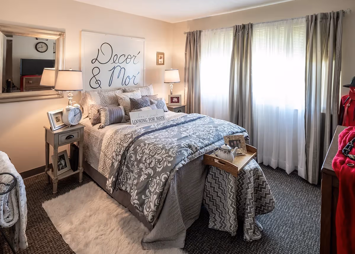 A cozy bedroom with a neatly made bed featuring patterned gray and white bedding and multiple pillows. There are two bedside tables with lamps, picture frames, and a clock. A large window with sheer and heavy curtains lets in natural light. A decorative tray with a teapot and photo frame sits on a small table at the foot of the bed. A mirror hangs on the wall above one bedside table, and a piece of wall art with the words 'Decor & Mor' is displayed above the bed.
