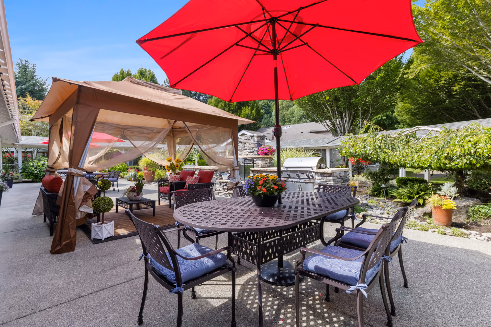 Outdoor patio area with a large red umbrella shading a metal table and four chairs with blue cushions. Nearby is a brown canopy tent with sheer curtains, containing cushioned seating and a coffee table. The area is surrounded by greenery, potted plants, and trees, with a stone grill in the background.