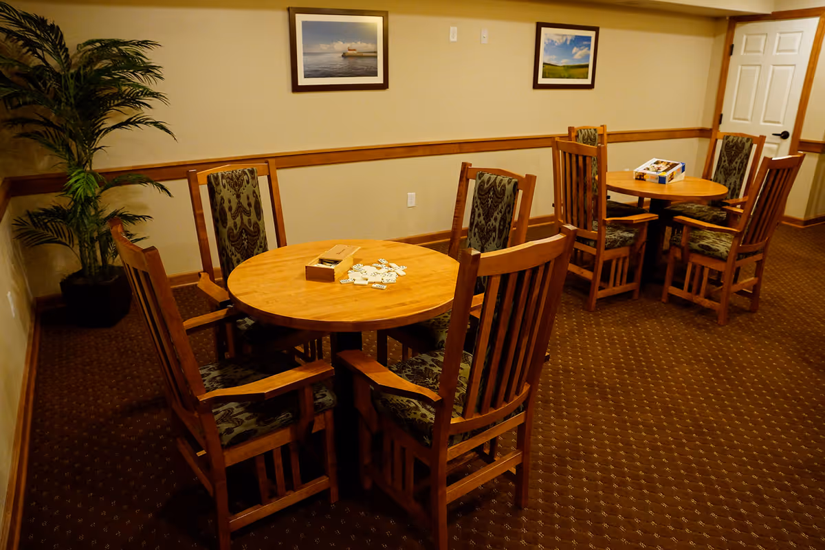 A cozy room with two round wooden tables, each surrounded by four wooden chairs with patterned cushions. One table has a domino game set on it, and the other has a puzzle box. The room has beige walls with wooden trim, two framed pictures hanging on the wall, a potted plant in the corner, and a closed white door.