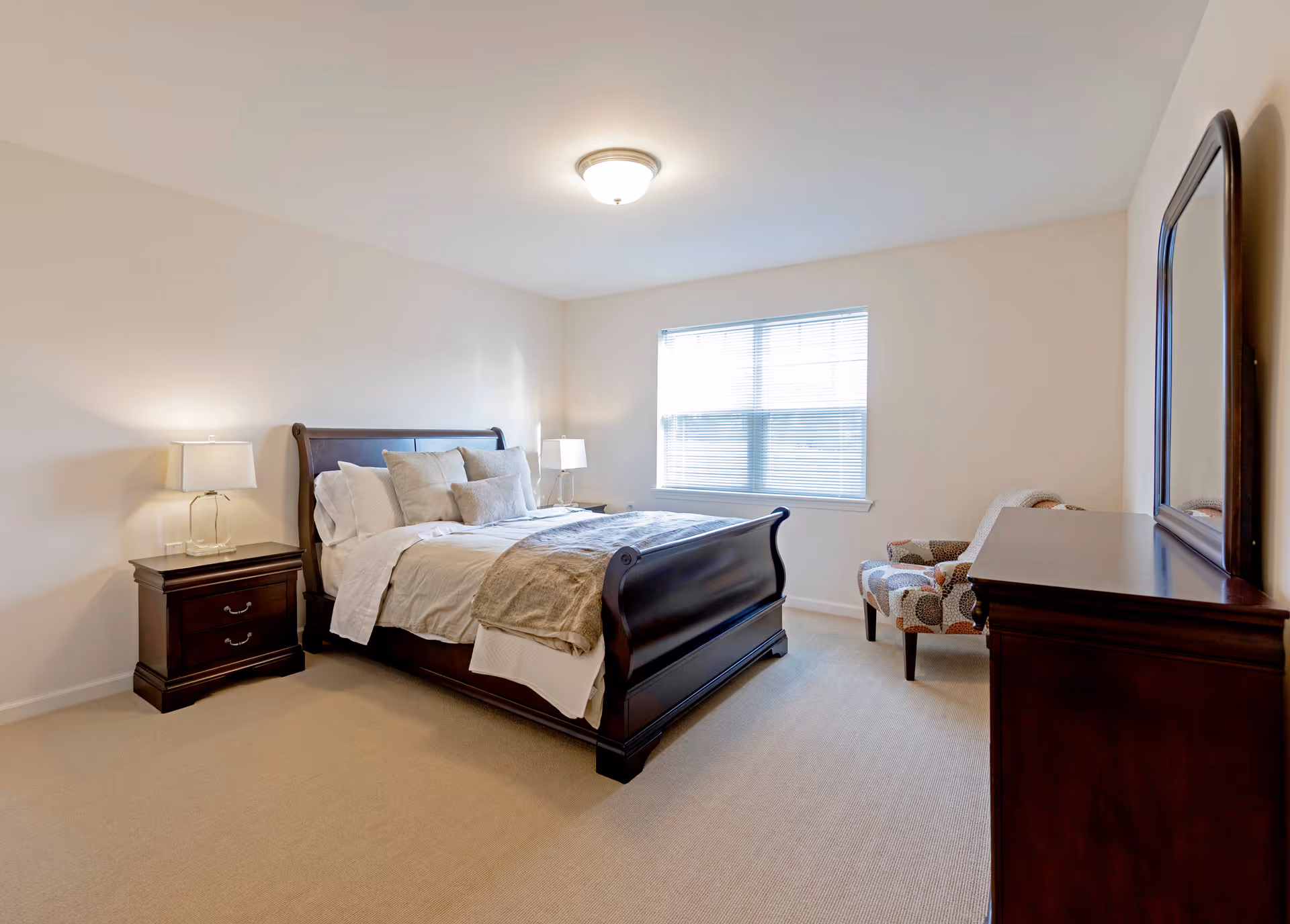 A bright bedroom with a large wooden bed featuring white and beige bedding and multiple pillows. There are two matching wooden nightstands with lamps on either side of the bed. A window with blinds lets in natural light. A patterned armchair is placed near a wooden dresser with a large mirror above it. The walls are painted light beige and the floor is carpeted.