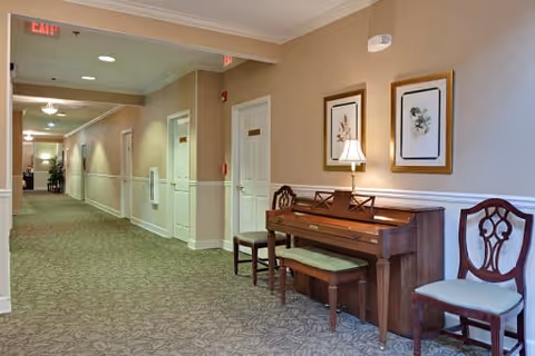 A carpeted hallway in an assisted living facility with beige walls and white trim. Along the right wall, there is a wooden piano with a lamp on top, a cushioned bench, and two wooden chairs with upholstered seats. Two framed botanical prints hang above the piano. Several doors line the hallway, and exit signs are visible on the ceiling.