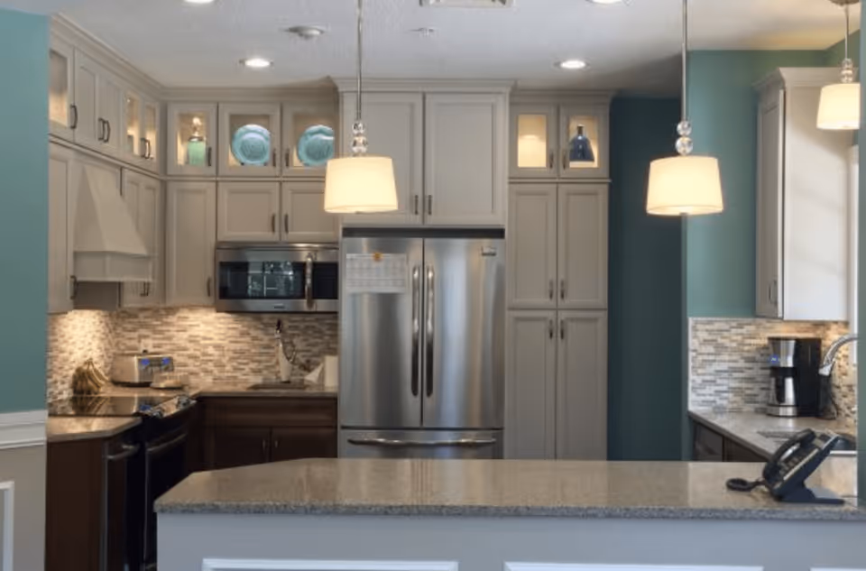 Modern kitchen with light gray cabinets, stainless steel refrigerator and microwave, granite countertops, two pendant lights hanging over a kitchen island, and a coffee maker on the right counter.