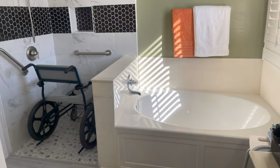 Bathroom with a white bathtub and a shower area equipped with grab bars and a wheelchair. Two towels, one orange and one white, hang on the wall above the bathtub. Sunlight streams through window blinds, casting shadows on the bathtub and wall.