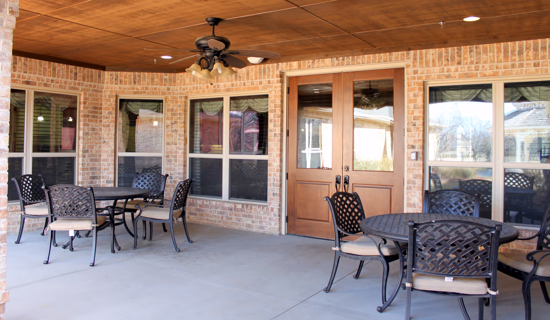 Covered outdoor patio area with brick walls, wooden ceiling, ceiling fan with lights, two round metal tables with cushioned chairs, and large wooden double doors leading inside.