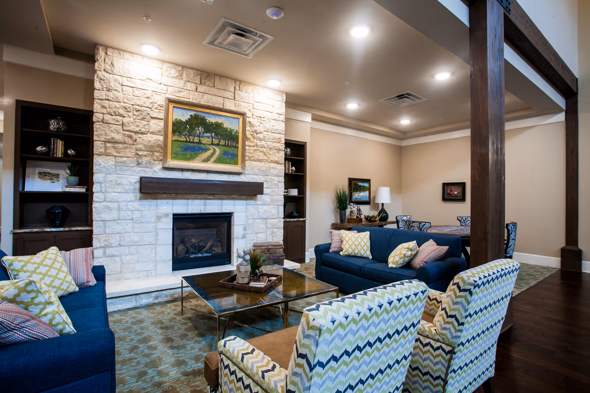 Communal living room with a stone fireplace, blue sofas, patterned armchairs, and a glass coffee table.