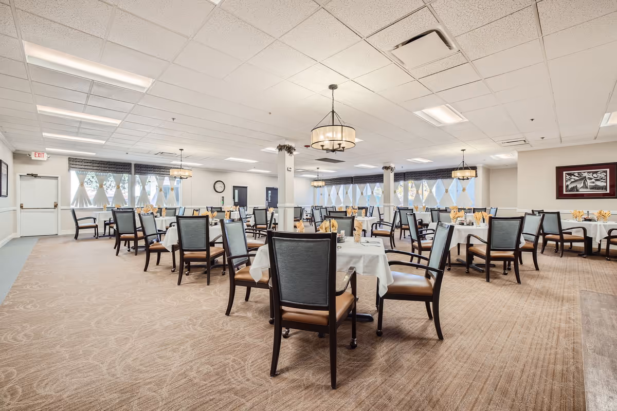 A spacious dining room in Winslow Court Assisted & Senior Living with multiple round tables covered with white tablecloths, each set with napkins and glassware. The room features carpeted floors, large windows with decorative curtains, and ceiling lights with hanging chandeliers. Chairs with cushioned seats and backs surround the tables.