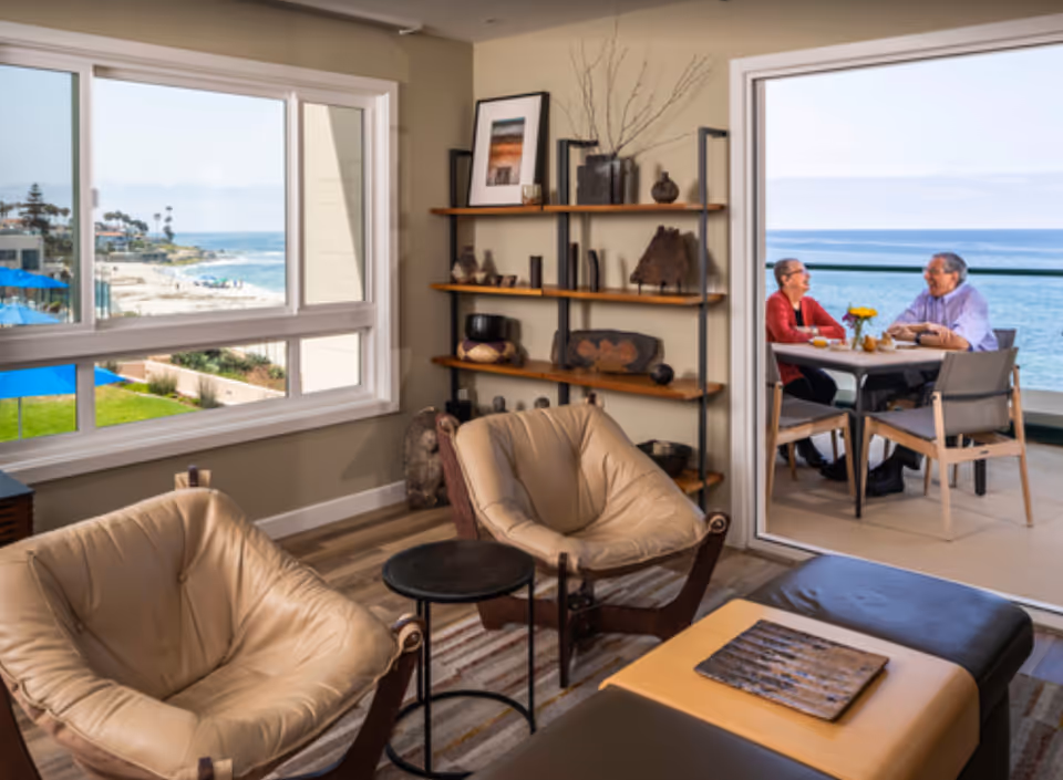 A bright living room with leather chairs and shelving that opens to a balcony where two people sit at a table overlooking the ocean.