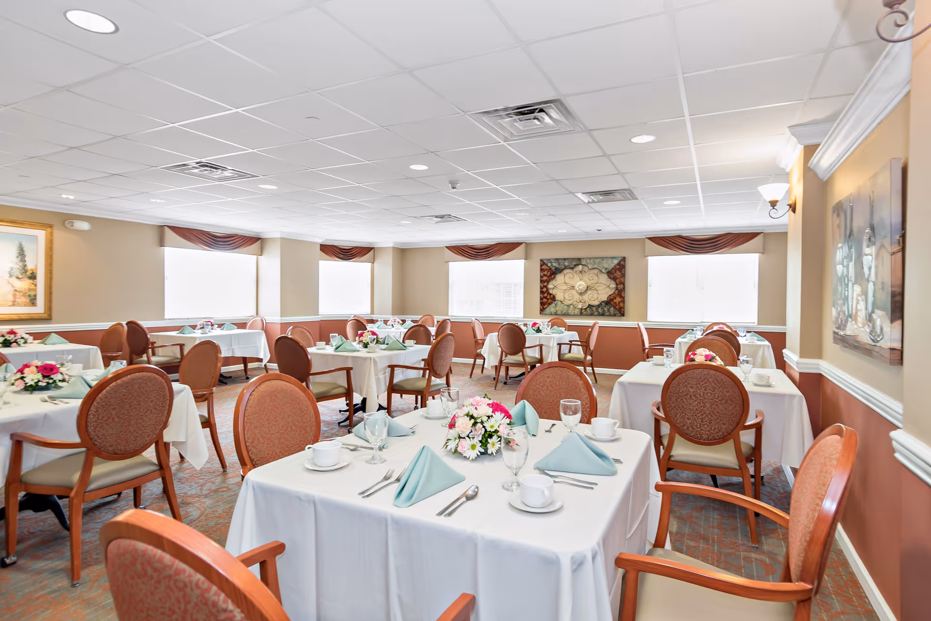 A bright dining room with multiple round tables covered in white tablecloths, each set with plates, cups, silverware, folded green napkins, and floral centerpieces. The room has beige walls with white trim, several large windows with valances, and framed artwork on the walls. Upholstered chairs with wooden frames surround the tables.