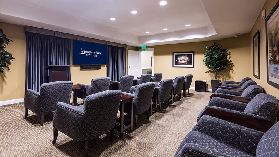 A small theater or presentation room with multiple rows of cushioned armchairs facing a wall-mounted screen displaying the Dougherty Ferry Assisted Living and Memory Care logo. The room has beige walls, carpeted floor, framed pictures on the walls, a podium, and a potted plant in the corner.