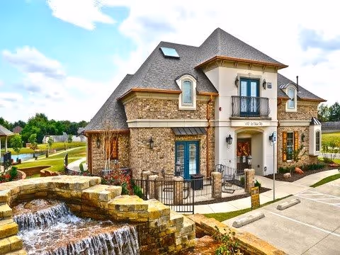A large, elegant building with stone and stucco exterior, featuring a balcony above the entrance and surrounded by landscaped grounds with a cascading water feature in the foreground under a partly cloudy sky.