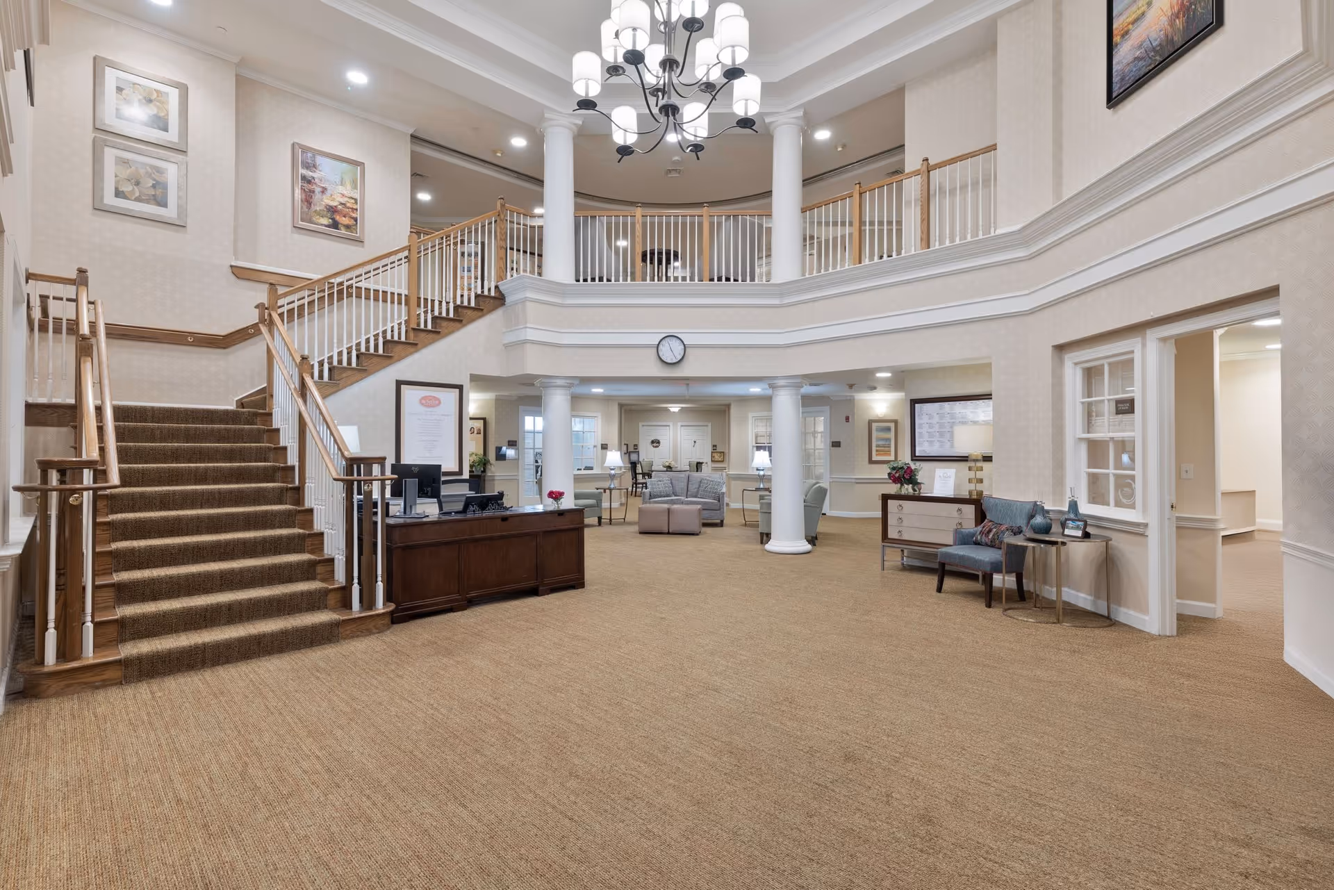 Spacious and well-lit interior lobby area of a senior living facility with a large staircase on the left, a chandelier hanging from the ceiling, a reception desk, seating area with sofas and chairs, decorative columns, framed artwork on the walls, and a clock above the seating area.