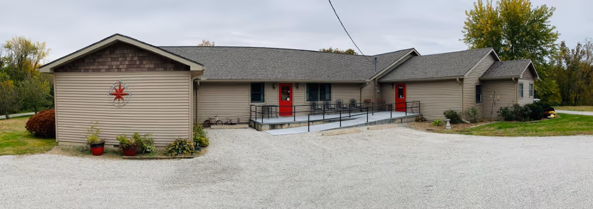 Exterior view of a single-story residential care center building with beige siding and a gray roof. The building features two red doors, a wheelchair ramp with black railings, and some landscaping with plants and bushes. The sky is overcast and there is a gravel driveway in front of the building.