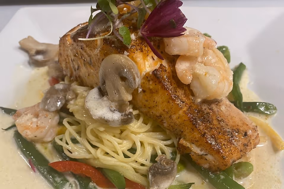 A plated dish featuring a grilled salmon fillet topped with shrimp and microgreens, served over a bed of pasta with sliced mushrooms, green beans, and red bell peppers in a creamy sauce.