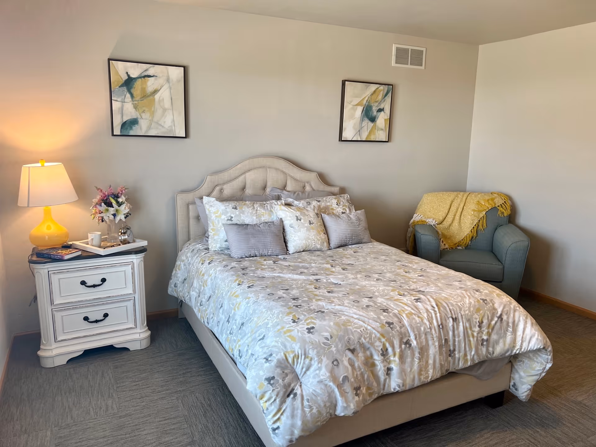 A neatly made bed with a floral patterned comforter and multiple pillows in a bedroom. To the left of the bed is a white nightstand with two drawers, a yellow lamp, a small flower arrangement, a cup, and some books. On the right side of the room is a blue armchair with a yellow throw blanket draped over it. Two abstract paintings hang on the wall above the bed.