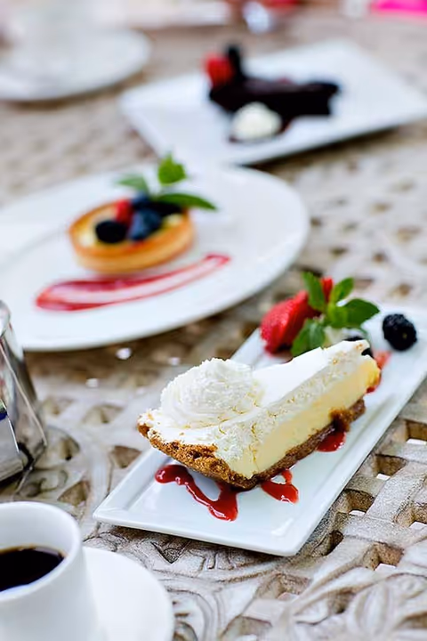A slice of cheesecake with whipped cream and berry garnish on a rectangular plate, with other plated desserts and a cup of coffee on a table.