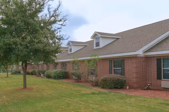 Exterior view of a single-story brick building with multiple windows and a sloped roof with dormer windows. The building is surrounded by a well-maintained lawn, bushes, and trees under a partly cloudy sky.