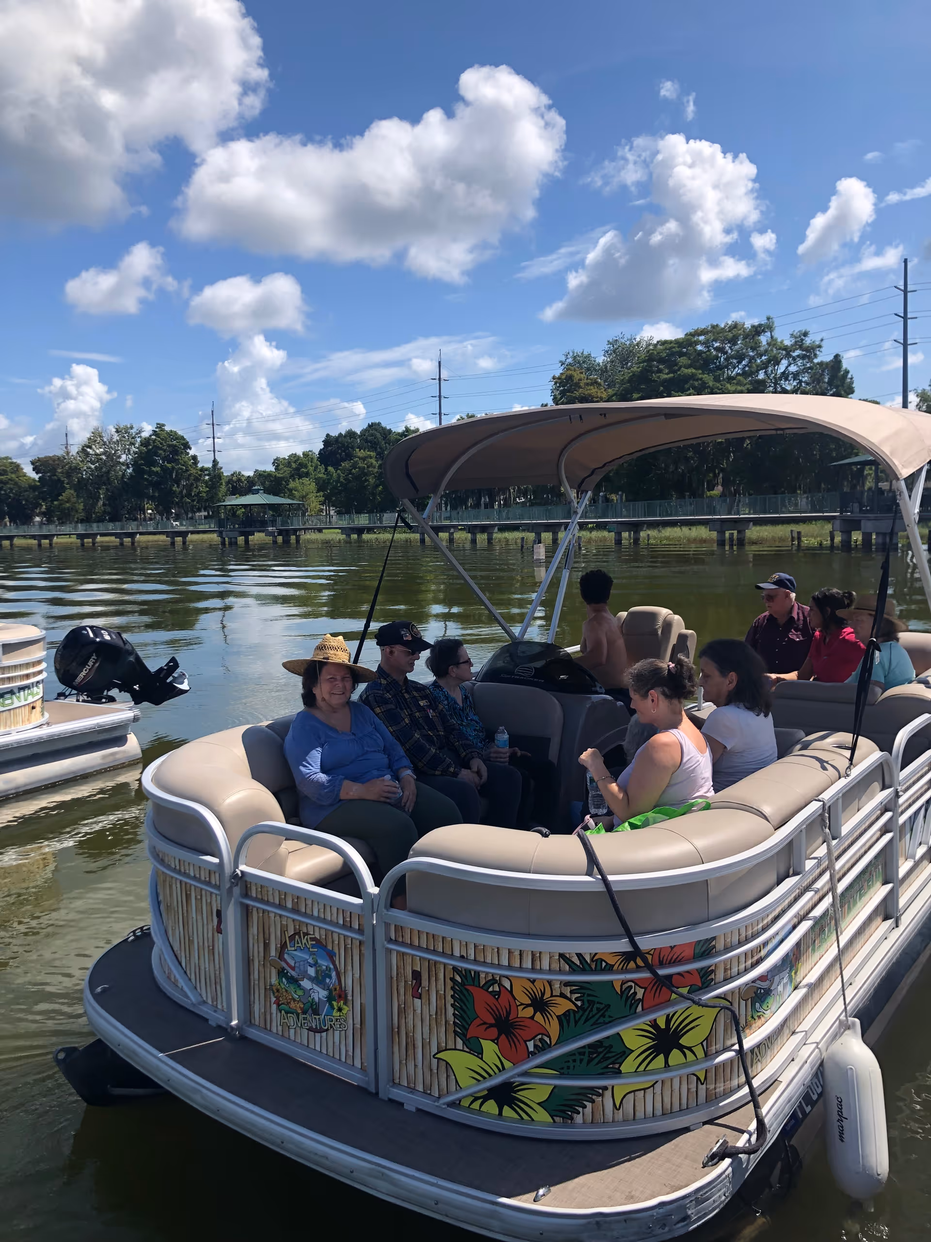 A group of people sitting and relaxing on a pontoon boat with a canopy on a calm lake under a partly cloudy blue sky. The boat has colorful floral decorations on the side and is surrounded by water with trees and a dock in the background.
