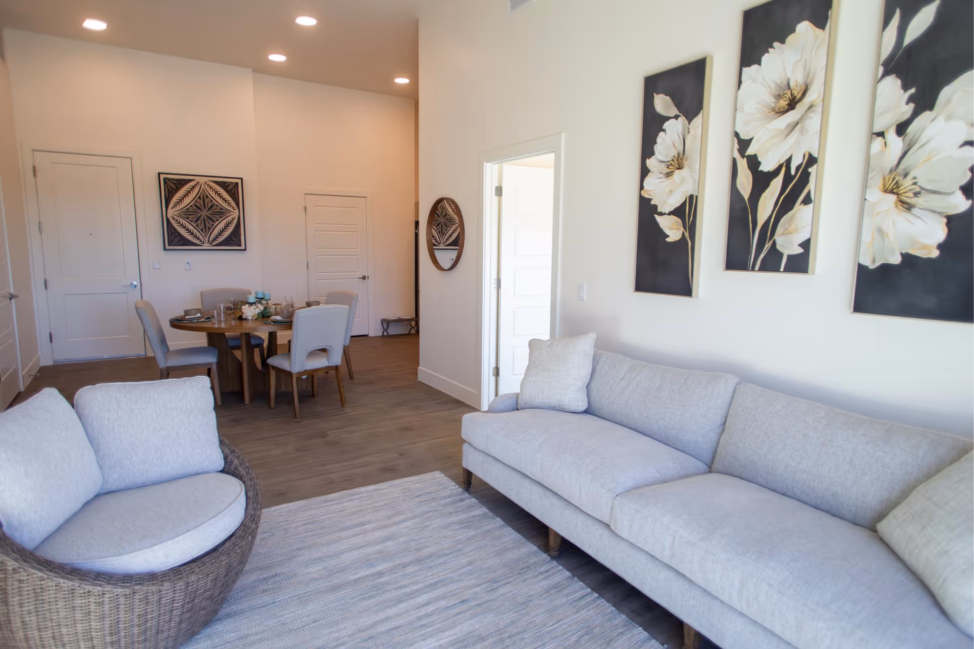 A bright and modern living room area with a light gray sofa and a round wicker chair with cushions. The walls are decorated with three large floral paintings. In the background, there is a dining area with a round wooden table and four upholstered chairs. The floor is wooden, and there is a light-colored rug in front of the sofa.