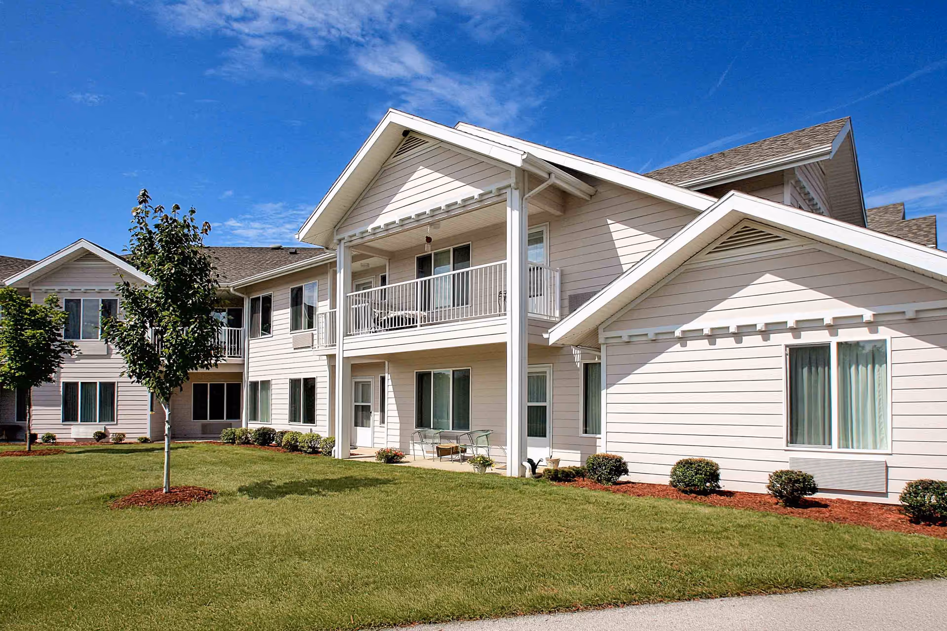 Two-story beige senior living building with balconies, manicured lawn and small trees under a blue sky.