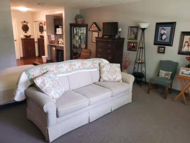 A cozy living room area with a light gray sofa adorned with two patterned pillows and a floral quilt draped over the back. Behind the sofa, there is a dark wooden dresser with a small TV on top and a glass display cabinet. The walls are decorated with framed family photos and a tall floor lamp stands beside a green upholstered chair. A small wooden side table with a plant is also visible. In the background, a kitchen area with a microwave and other appliances can be seen.
