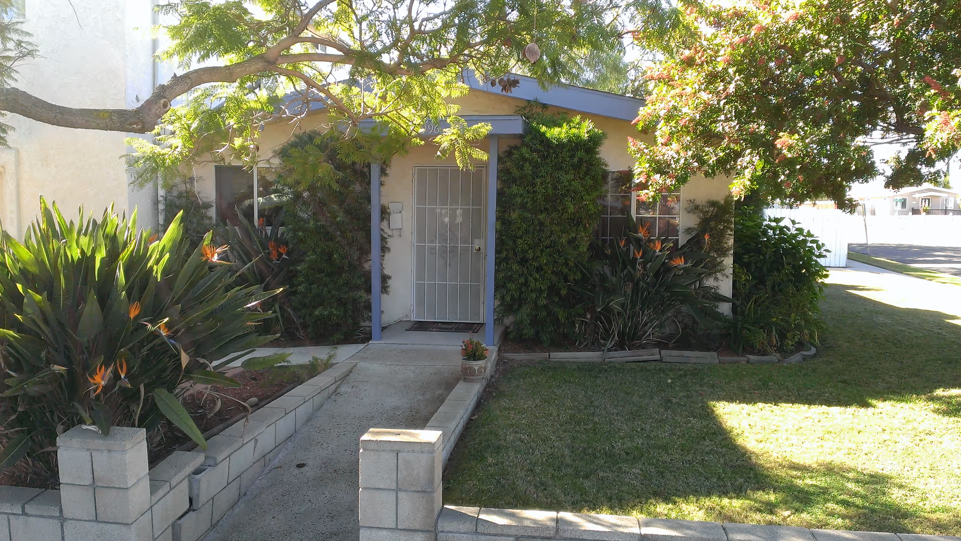 Front exterior view of a single-story house with a small porch, a metal security door, and windows partially covered by green bushes and plants. There is a concrete walkway leading to the entrance, bordered by a low brick wall and landscaped with flowering plants and trees providing shade.
