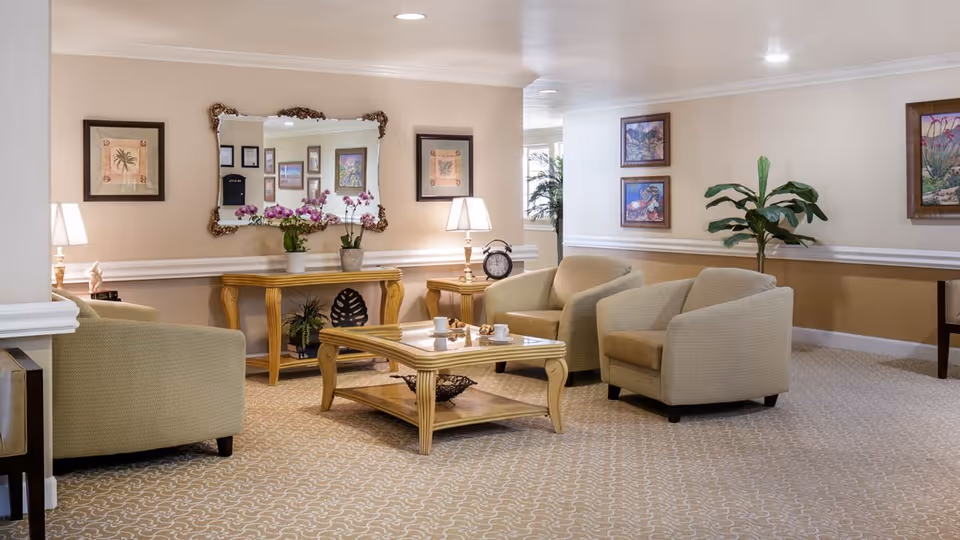 A cozy living room area with beige armchairs arranged around a wooden coffee table with cups on it. Behind the chairs is a wooden console table with potted orchids and a decorative mirror above it. The walls are adorned with framed artwork, and there is a potted plant in the corner. The room has soft lighting from table lamps and recessed ceiling lights.