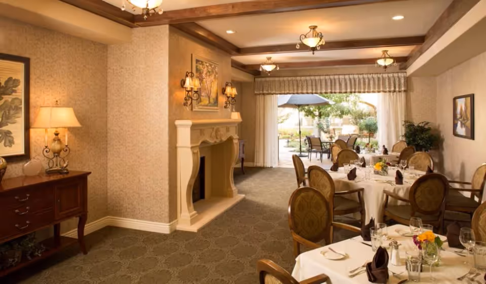A warmly lit dining room with round tables covered in white tablecloths, set with glasses, napkins, and floral centerpieces. The room features a decorative fireplace, wall sconces, framed artwork, and a side table with a lamp. Large windows with curtains open to an outdoor patio area with tables, chairs, and an umbrella surrounded by greenery.
