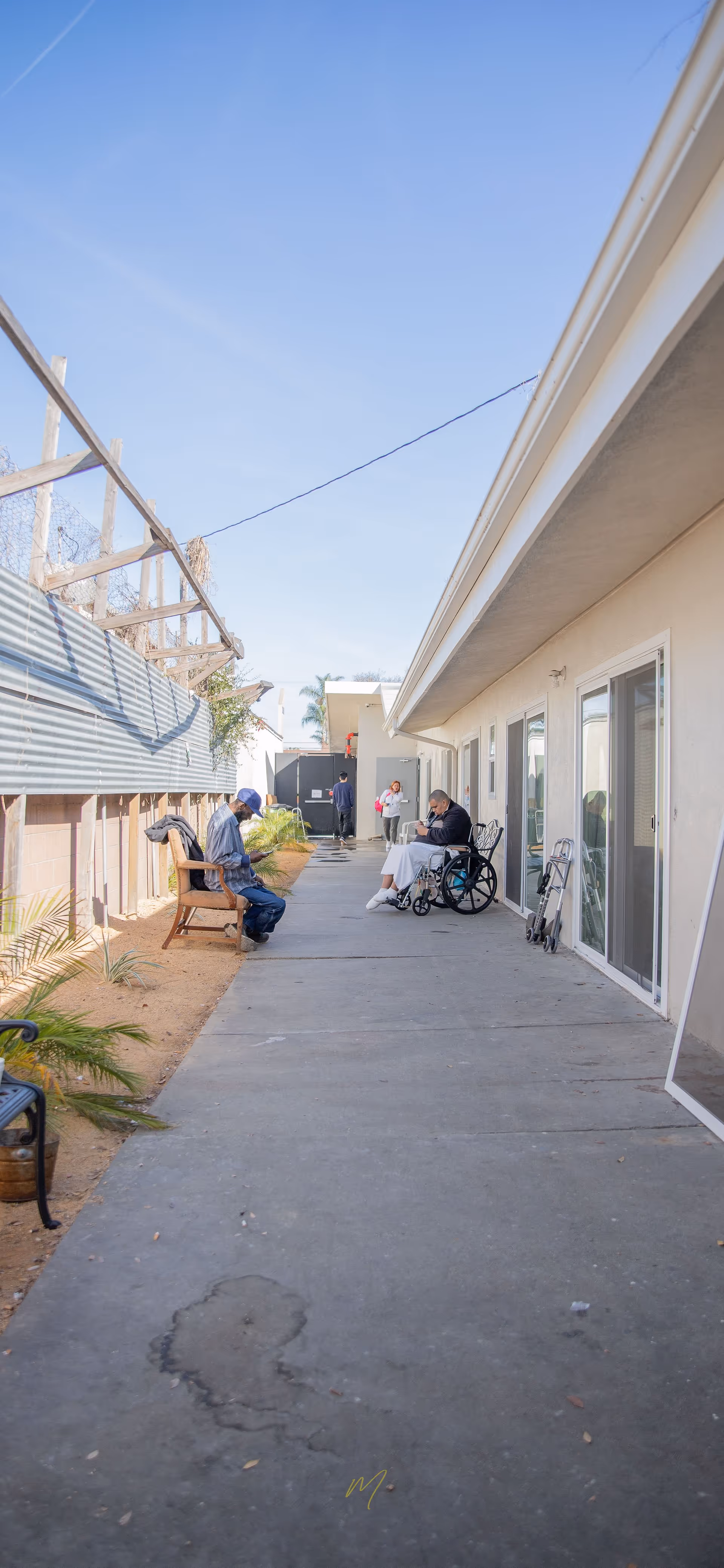 Outdoor walkway area at Highland Manor Assisted Living with a man sitting on a bench on the left and another man in a wheelchair on the right near the building. Two other people are walking in the background. The sky is clear and blue.
