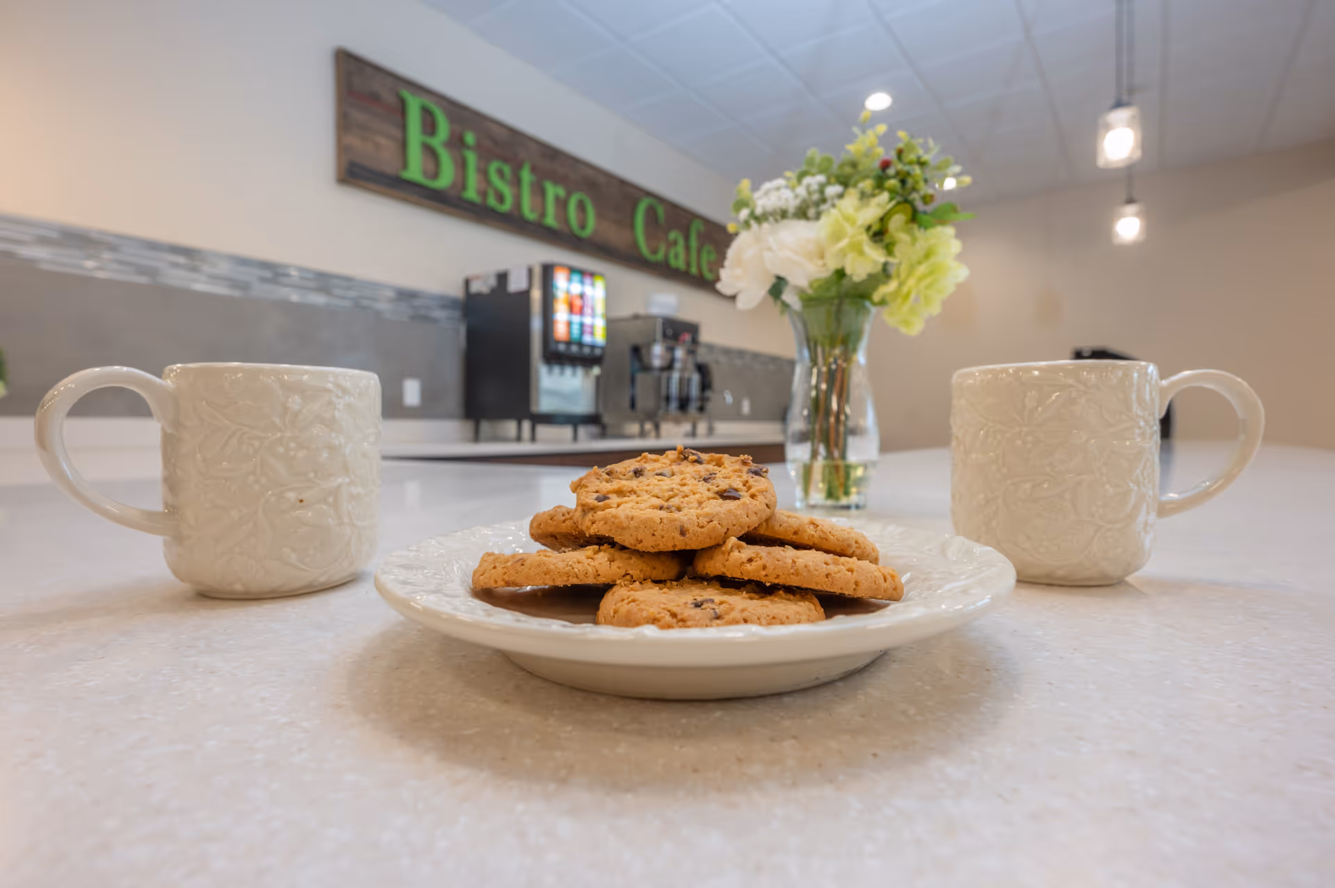 Close-up of a plate with chocolate chip cookies on a countertop, flanked by two white ceramic mugs with floral patterns. In the background, a vase with a bouquet of white and green flowers is visible, along with a beverage dispenser and a coffee machine under a sign that reads 'Bistro Cafe'.