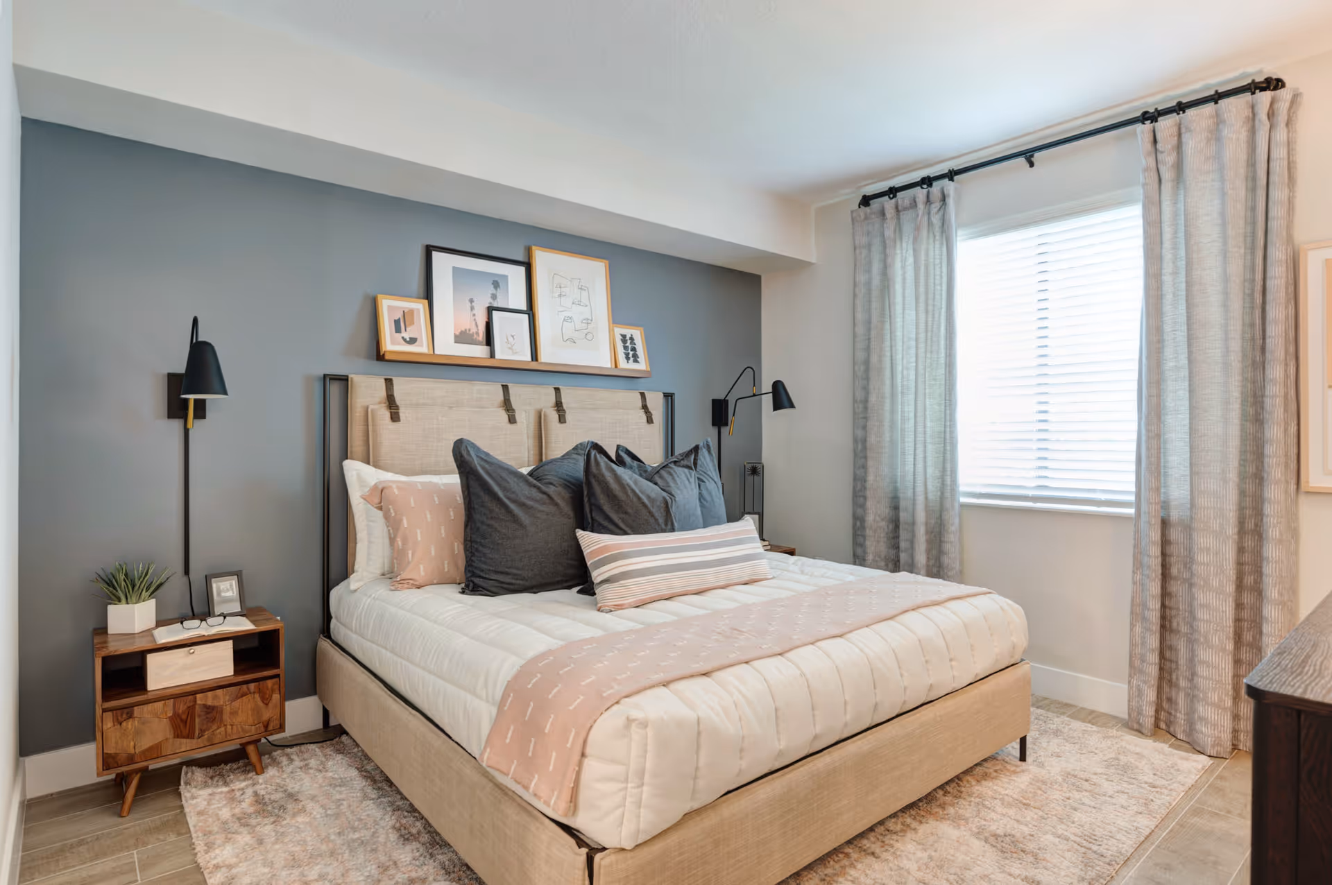 A modern bedroom with a beige upholstered bed featuring multiple pillows in dark gray and light pink. The bed is against a gray accent wall with a shelf holding framed artwork. There are two black wall-mounted reading lamps on either side of the bed. A wooden nightstand with a small plant, a book, and glasses is on the left side. Light gray curtains frame a window with white blinds, and a light-colored rug covers the floor.
