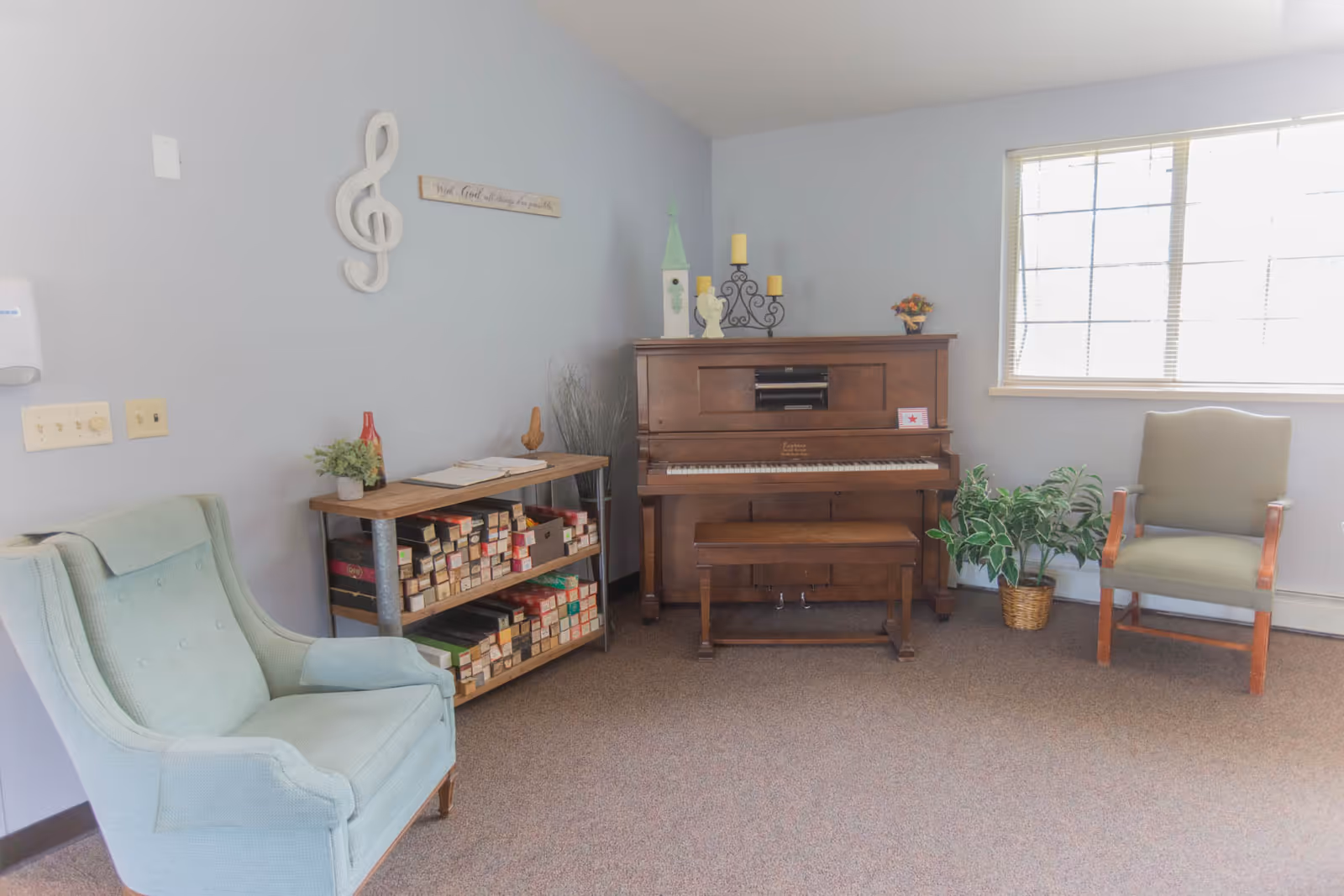 A cozy room with a wooden upright piano against a light gray wall, flanked by two armchairs, one light blue and one green. A small wooden shelf with various boxes and a plant sits to the left of the piano. A window with blinds lets in natural light on the right side. Decorative items including a treble clef wall art, candles, and a small plant add a homey touch.