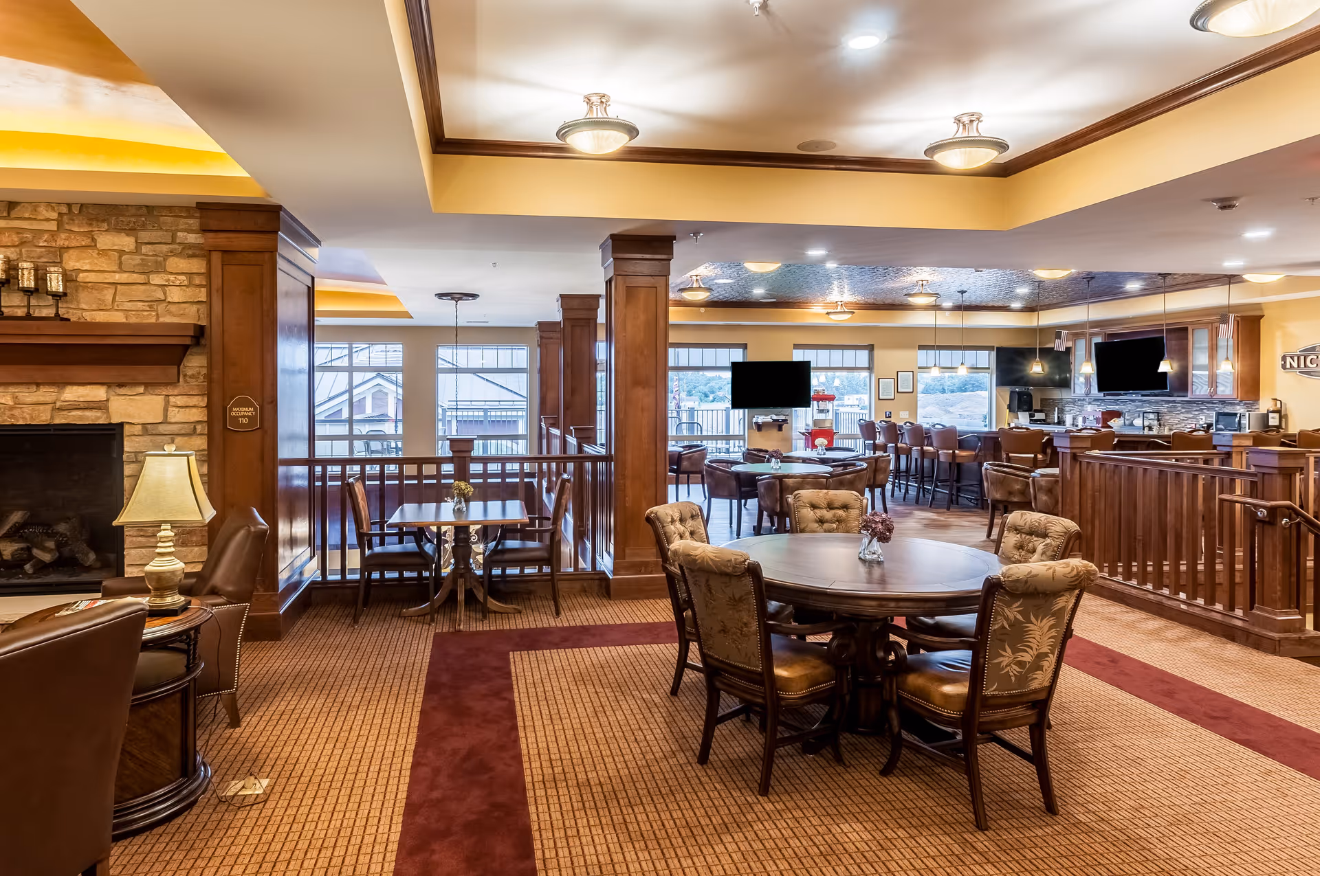 A spacious, warmly lit common area in a senior living facility featuring multiple seating arrangements including round tables with upholstered chairs, a fireplace with a lamp on a side table, and a bar area with high chairs. Large windows provide natural light and views outside.