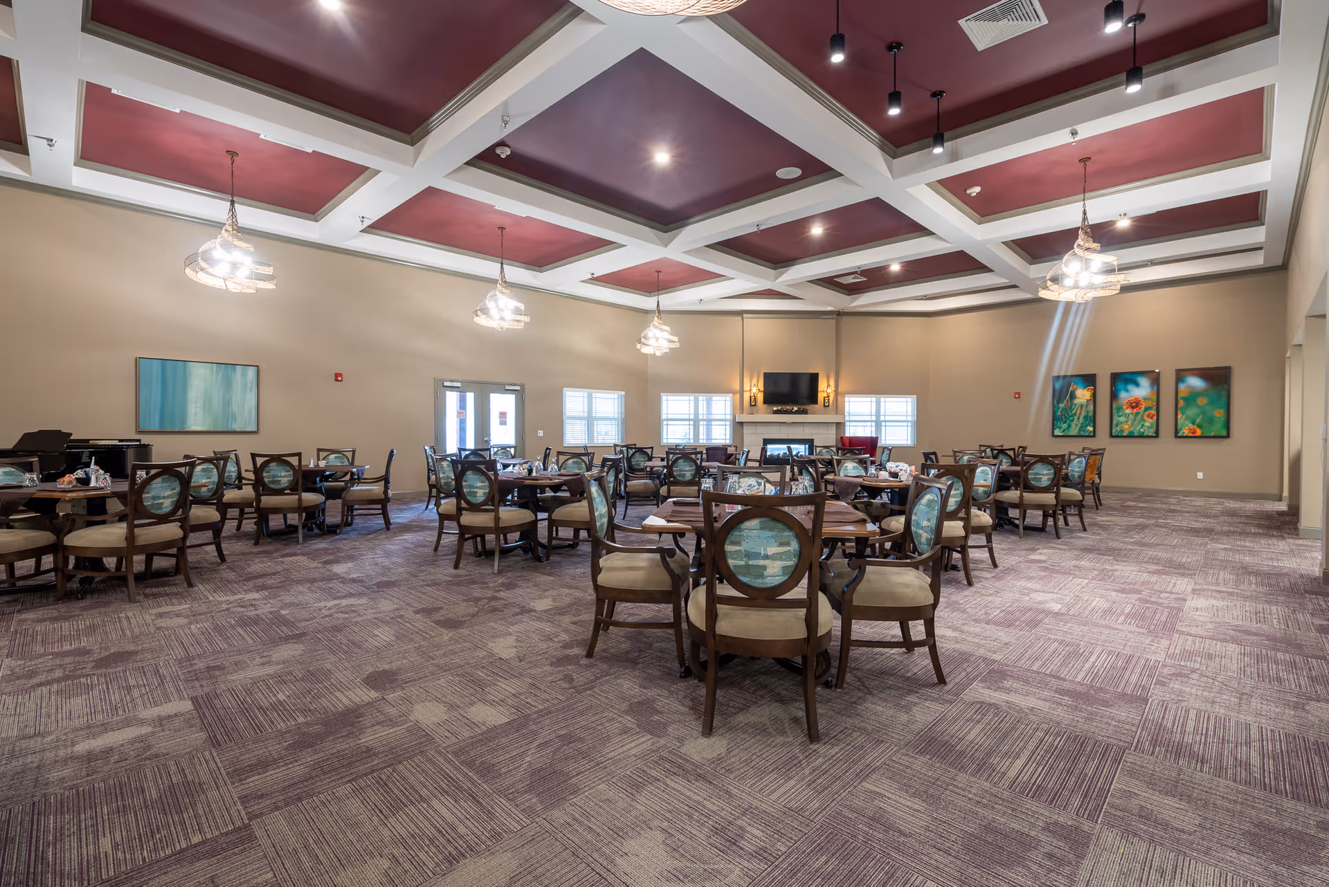 Spacious dining room with multiple round tables and upholstered chairs, a fireplace and TV at the far wall, and a coffered ceiling with pendant lights.