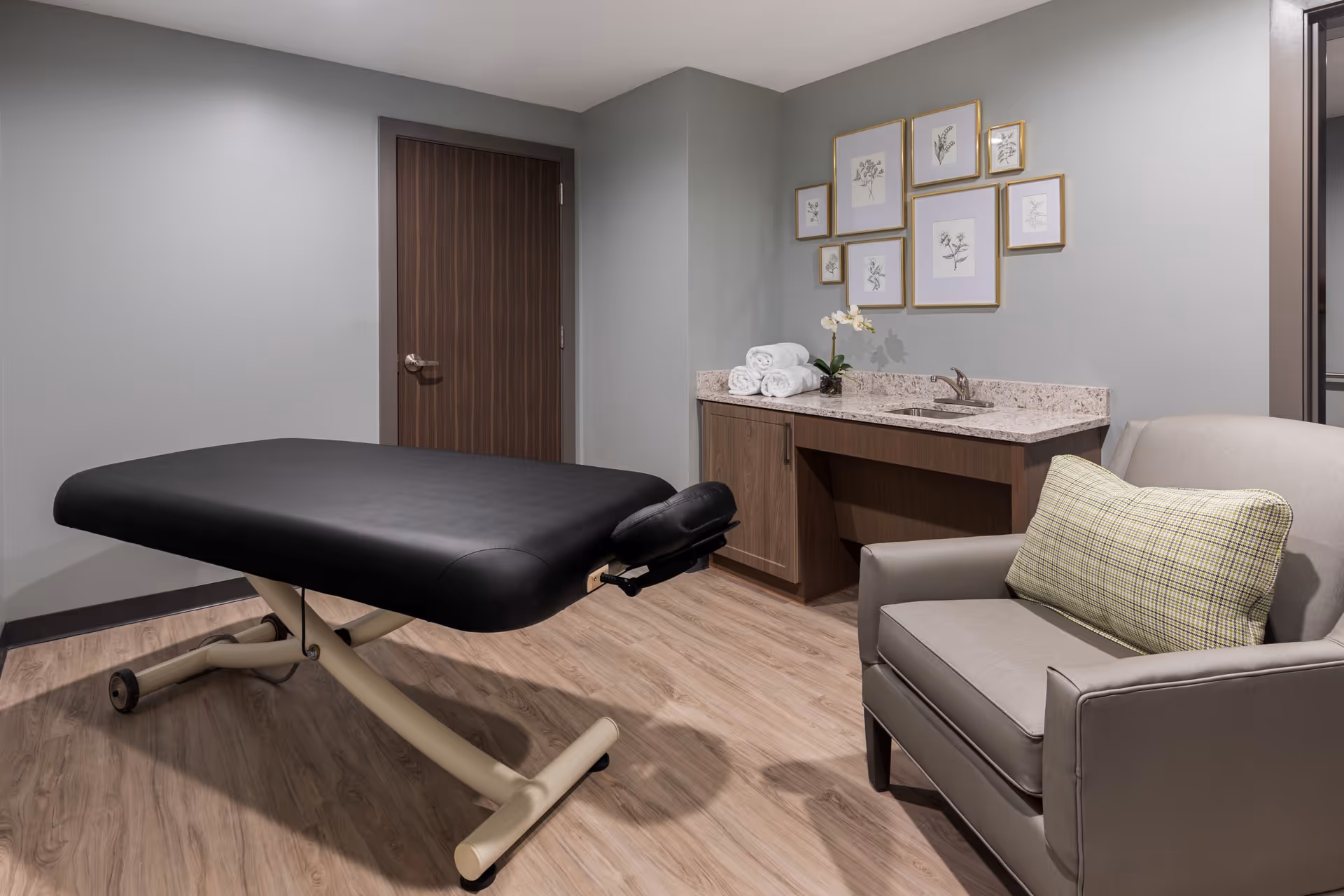 A therapy or massage room with a black padded treatment table, a gray armchair with a plaid cushion, a wooden cabinet with a granite countertop, a small sink, rolled white towels, a potted orchid, and framed botanical artwork on the wall.