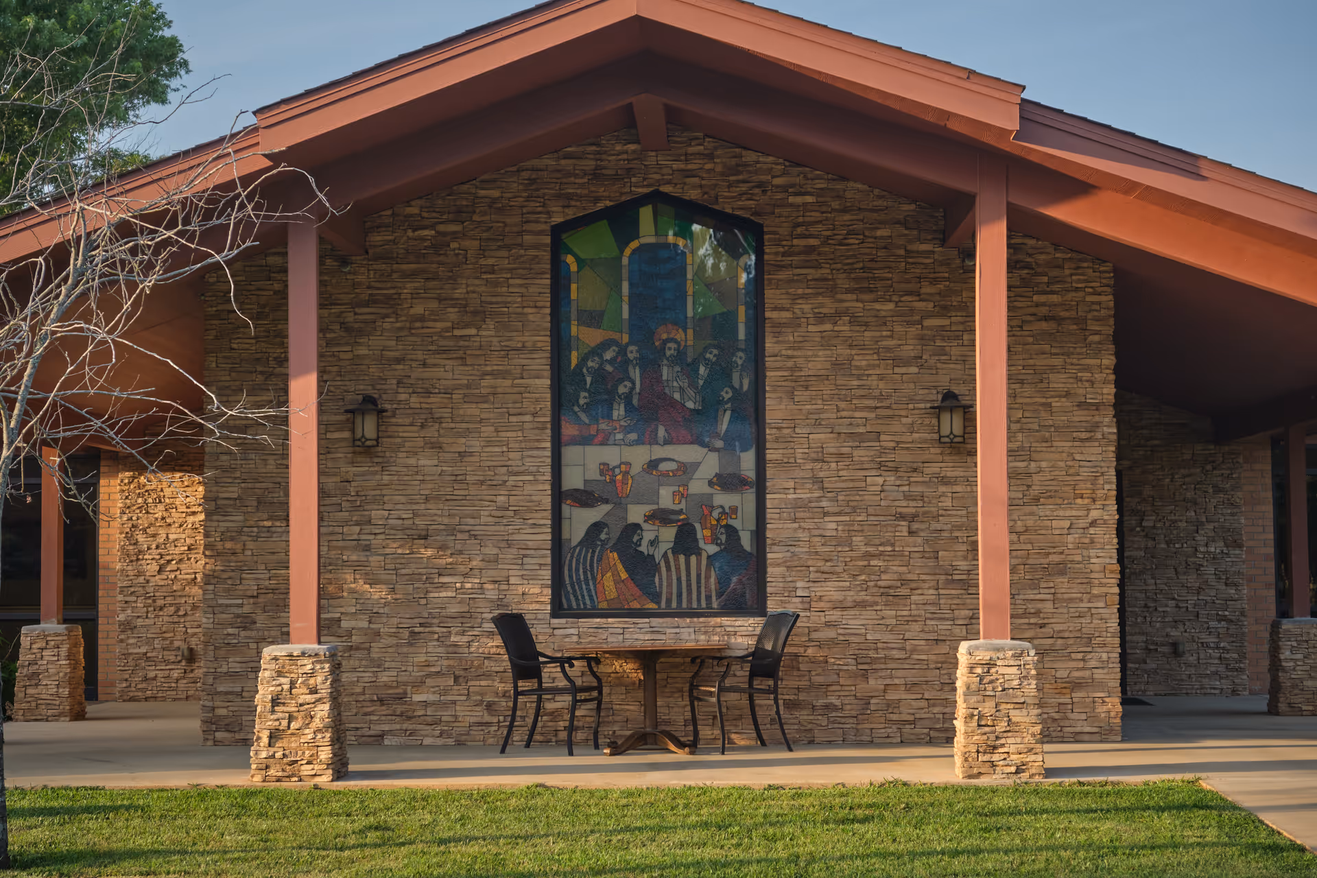 Outdoor view of a stone building with a peaked roof and reddish-brown beams. There is a large stained glass window depicting a religious scene in the center of the wall. Below the window is a small round table with two black chairs on a concrete patio. Green grass is visible in the foreground.