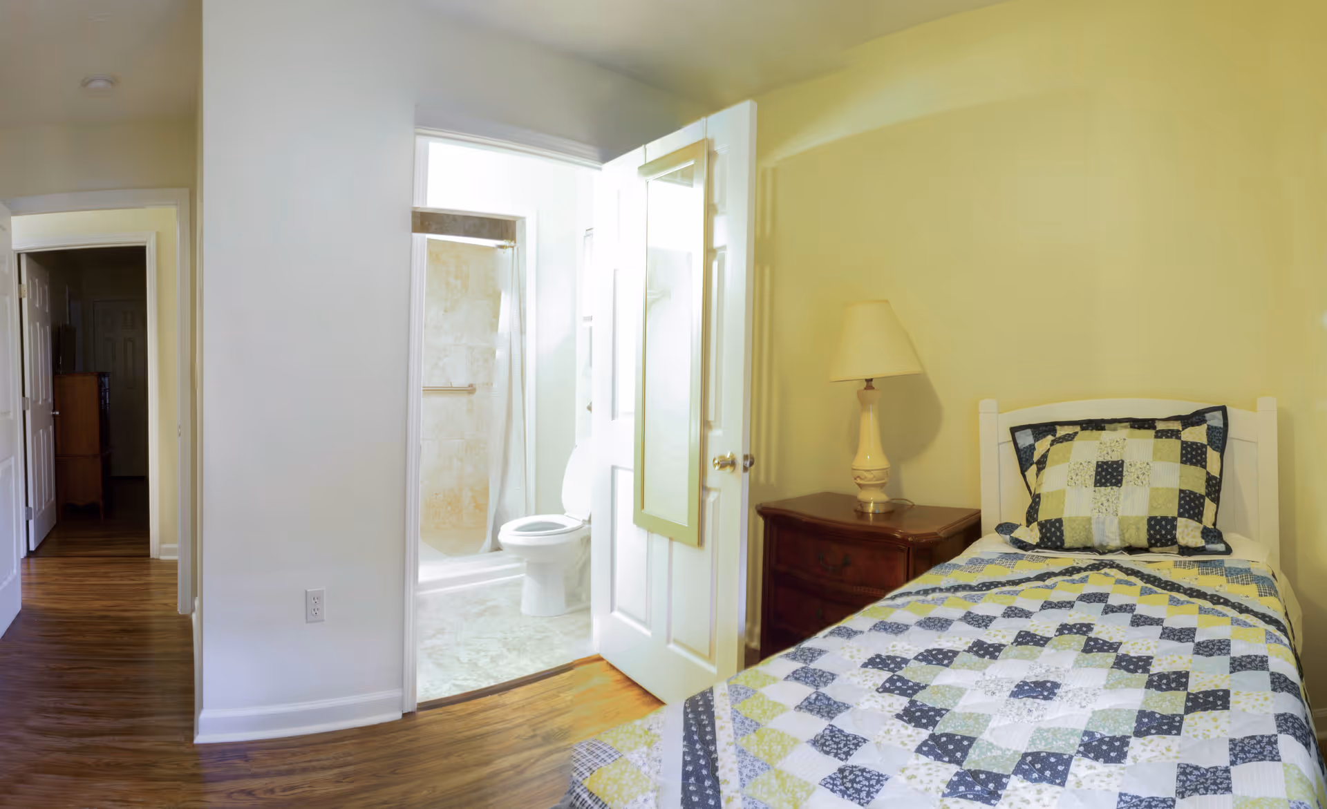 A small bedroom with a single bed covered in a patchwork quilt and a matching pillow. Next to the bed is a wooden nightstand with a lamp. The room has light yellow walls and wooden flooring. An open door reveals a bathroom with a toilet and a shower with tiled walls.