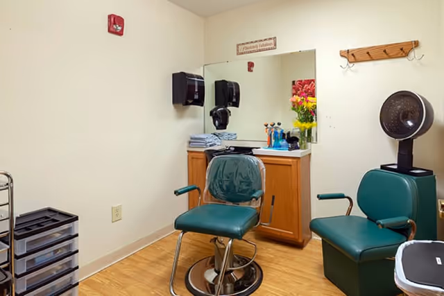 Small salon-style room with green barber chairs, a mirror and countertop with hair-care supplies and a hooded dryer.