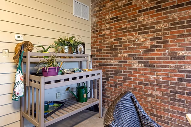 A small indoor gardening station with a wooden potting bench holding potted plants, gardening tools, and a watering can, set against a beige paneled wall and a red brick wall. A woven chair is partially visible in the foreground.