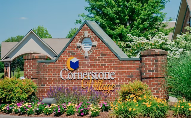 Brick entrance sign for Cornerstone Village surrounded by colorful flowers and greenery with trees and a building partially visible in the background under a clear blue sky.