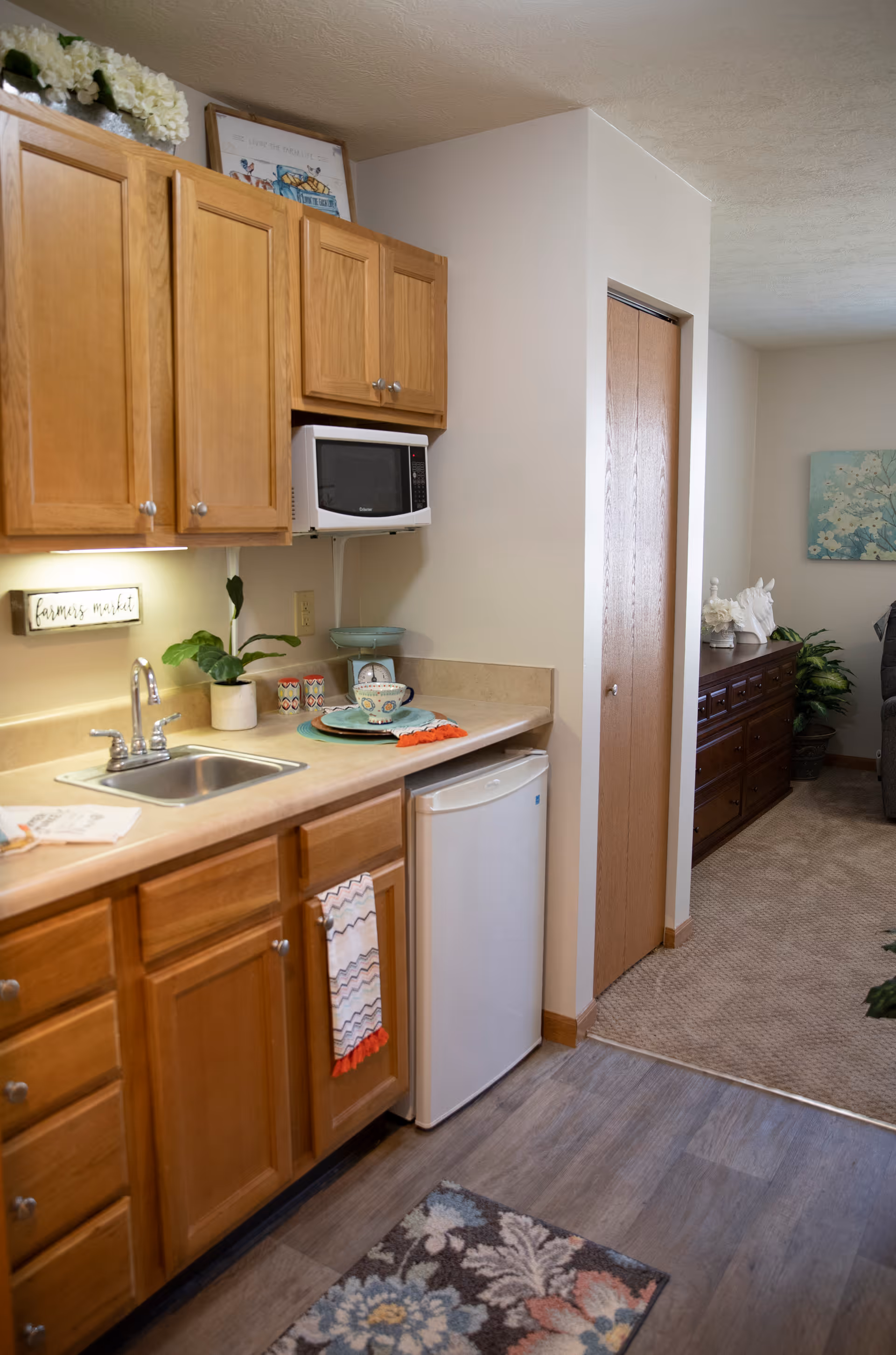 Small kitchenette with wooden cabinets, a sink, microwave, mini fridge and countertop decor opening into an adjacent living area.