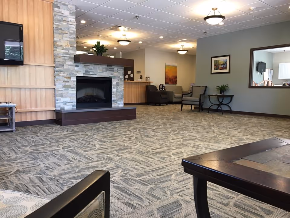 A spacious and well-lit common area with patterned carpet flooring, a stone fireplace, several chairs and small tables, a wall-mounted TV, and a reception desk in the background. The walls are painted in neutral tones with framed artwork and a window opening to another room.