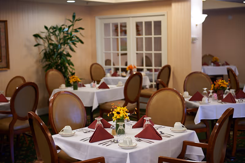 Dining room with multiple tables set with white tablecloths, folded burgundy napkins, place settings, and small floral centerpieces.