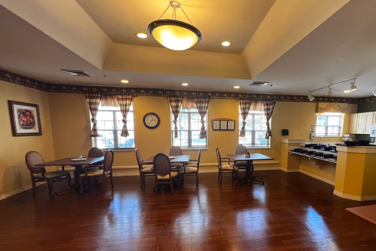 A dining room with three tables, each surrounded by chairs. The room has wooden flooring, yellow walls, and windows with plaid curtains letting in natural light. A clock and framed pictures hang on the walls. To the right, there is a kitchen area with multiple stove burners and cabinets.