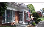Exterior view of a brick building with a white entrance door and large windows. There are American flags displayed near the entrance, potted plants, and a bench on the porch. The surrounding area includes a sidewalk, trimmed bushes, and trees with a neighboring house visible in the background.
