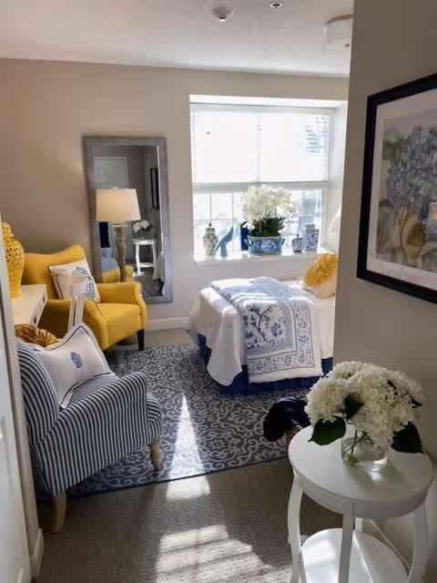A cozy bedroom with a single bed covered in white and blue patterned bedding. There is a large window with white blinds letting in natural light. A yellow armchair and a striped armchair are placed near the bed. A tall mirror stands against the wall next to a table lamp. Decorative vases and a potted plant are on the windowsill. A small round white table with a vase of white flowers is in the foreground. A framed floral artwork hangs on the wall.