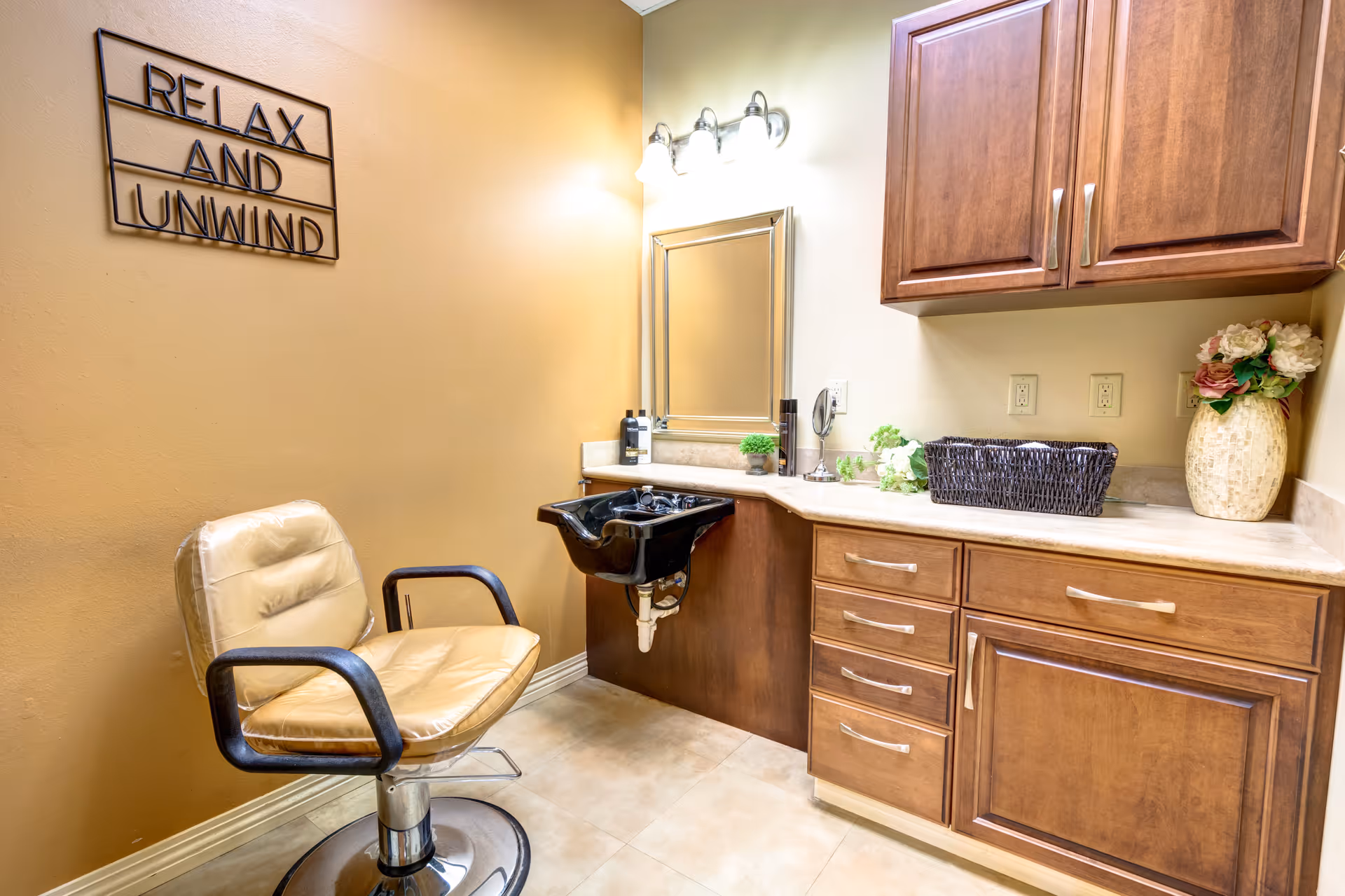 Small salon room with a beige styling chair, a black shampoo sink, wooden cabinets, and a 'Relax and Unwind' wall sign.