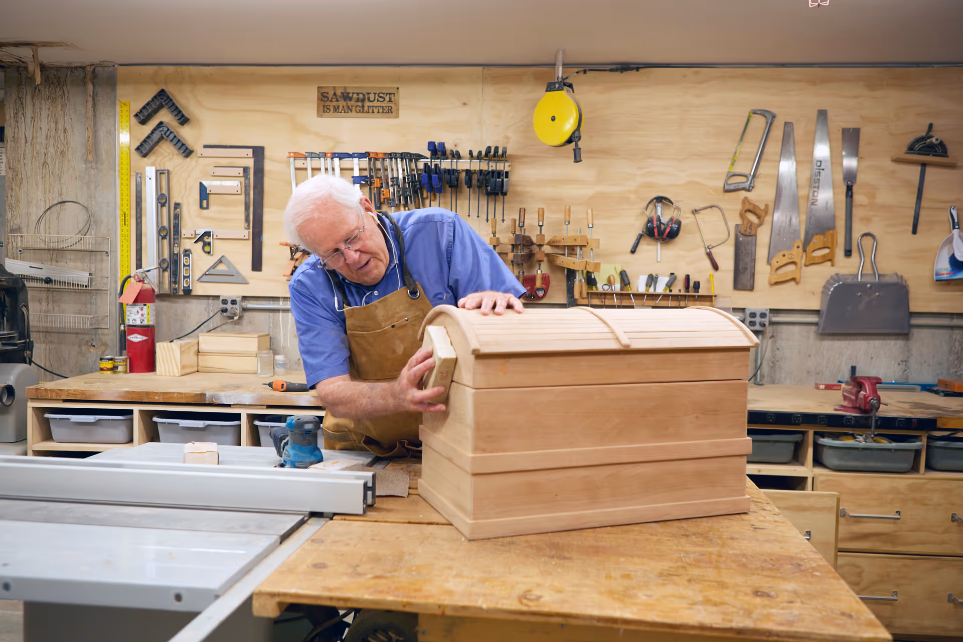 An elderly man wearing a blue shirt and brown apron is working on a wooden chest in a well-organized woodworking workshop. Various woodworking tools such as saws, clamps, and squares are neatly hung on a wooden pegboard behind him. A sign on the wall reads 'SAWDUST IS MAN GLITTER.'