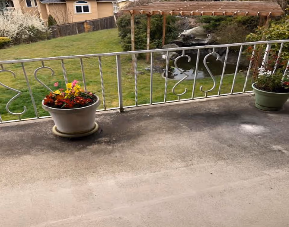 View from a concrete balcony with a decorative metal railing and potted flowers overlooking a grassy backyard with a pergola and pond.