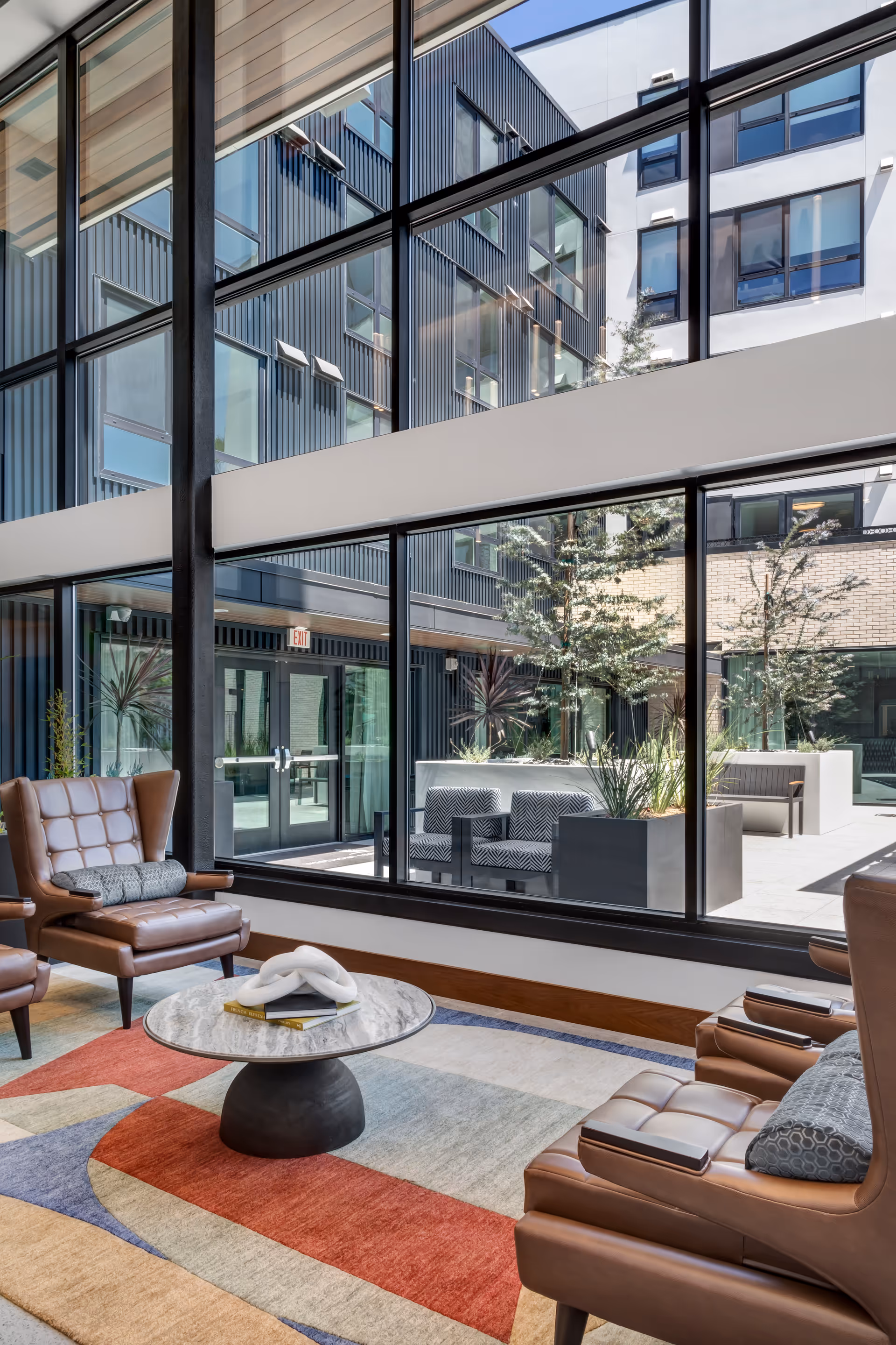 Modern indoor lounge area with large floor-to-ceiling windows overlooking an outdoor courtyard. The lounge features brown leather armchairs with cushions, a round marble coffee table on a colorful geometric rug, and visible plants both inside and outside.