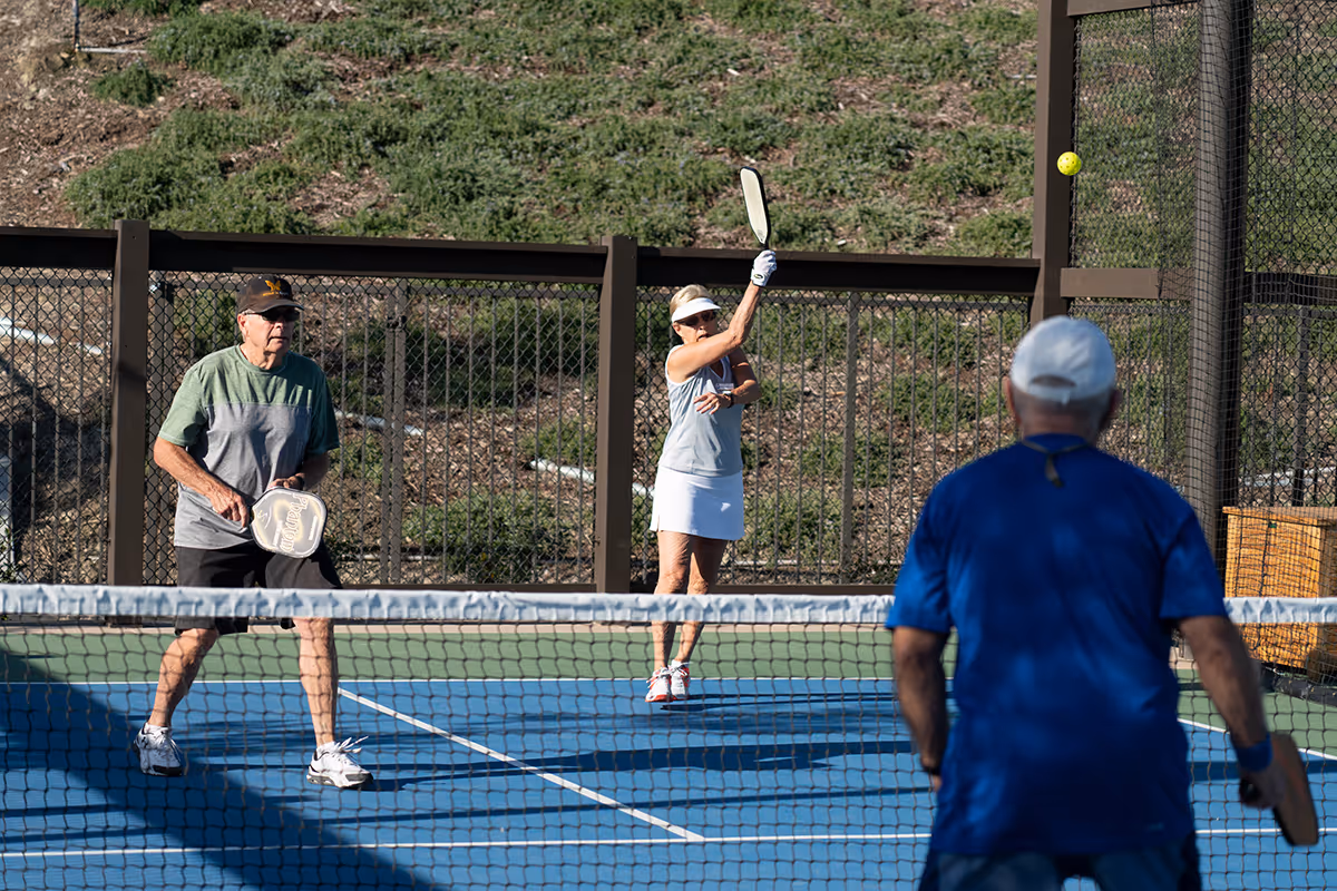Three senior adults playing pickleball on an outdoor court with a chain-link fence and hillside in the background. One woman is hitting the ball with a paddle while two men watch and prepare to play.